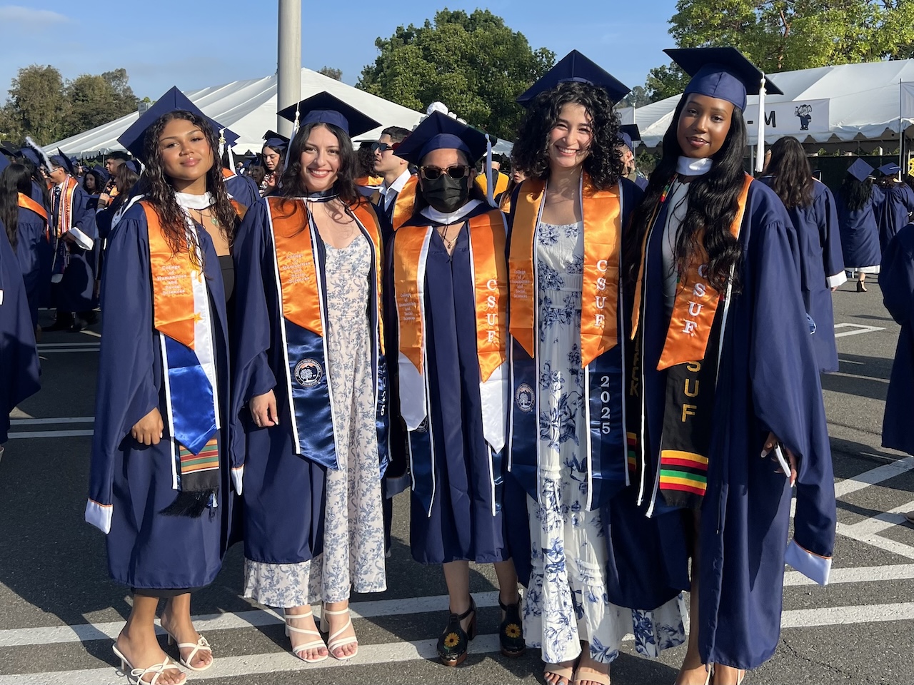 Five Master of Arts students posing together wearing blue graduation caps and robes with orange sashes