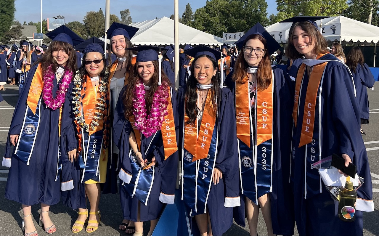 Seven Master of Science students posing together outside wearing blue graduation caps and robes with oranges sashes